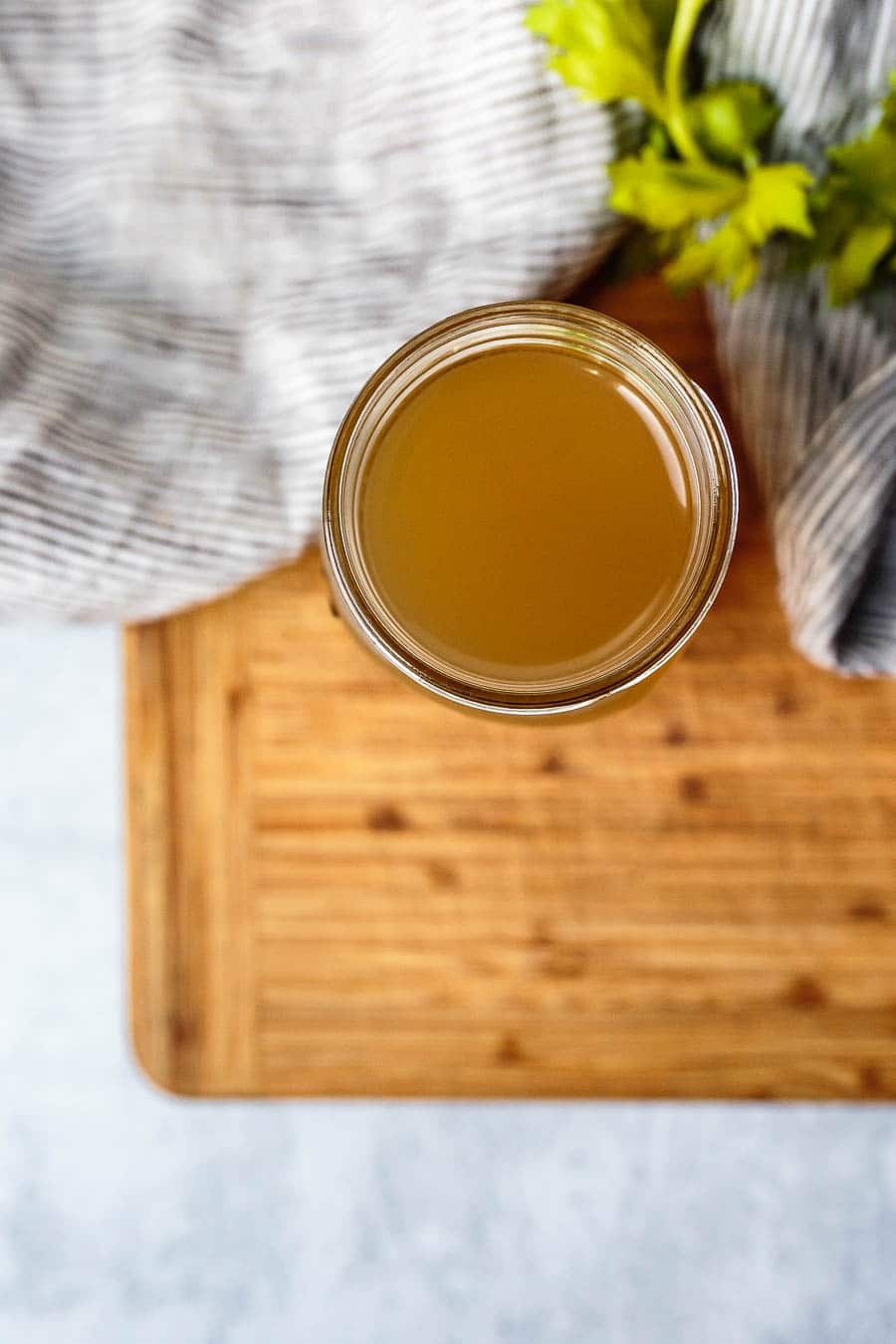 Aerial view of chicken bone broth in a mason jar. The jar doesn't have a lid on and is sitting on a wooden cutting board. There is also a dish cloth and herbs laying next to it.