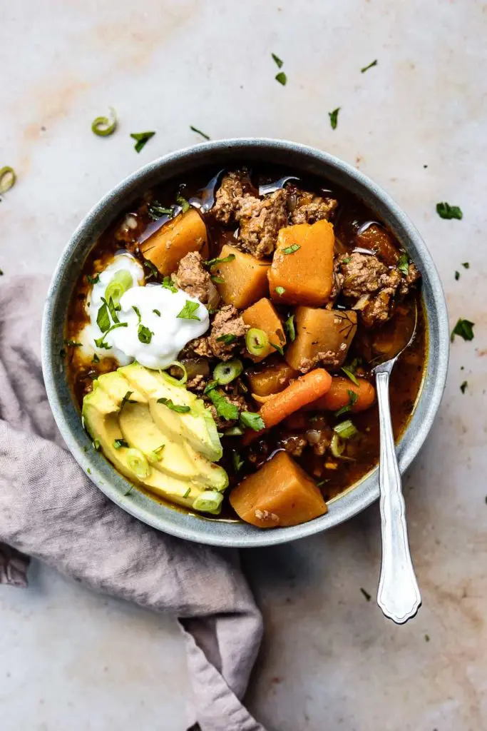 an overhead shot of a stoneware bowl of this Instant Pot Paleo Chili Recipe with sliced avocado, coconut yogurt, sliced green onions, and chopped parsley with a silver soup spoon and a cloth napkin | AIP chili chili with no tomatoes instant pot chili no beans