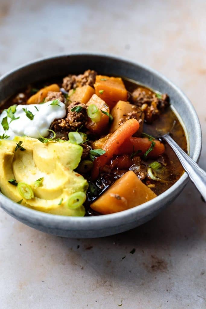 a stoneware bowl of this Instant Pot Paleo Chili Recipe with sliced avocado, coconut yogurt, sliced green onions, and chopped parsley with a silver soup spoon and a cloth napkin | AIP chili chili with no tomatoes instant pot chili no beans