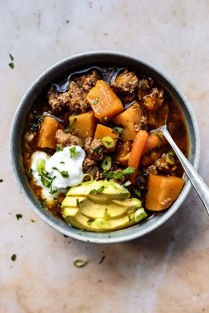 an overhead shot of a stoneware bowl of this Instant Pot Paleo Chili Recipe with sliced avocado, coconut yogurt, sliced green onions, and chopped parsley with a silver soup spoon and a cloth napkin | AIP chili chili with no tomatoes instant pot chili no beans