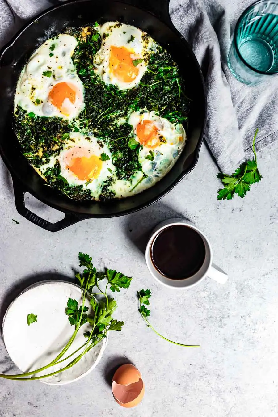 View from above of green breakfast shakshuka in a black skillet. Inside the skillet all the green veggies have wilted and the eggs can be seen very vibrant. The skillet is on a white table top next to a blue glass, a cup of coffee is also on the table, next to an empty egg shell and some fresh herbs.