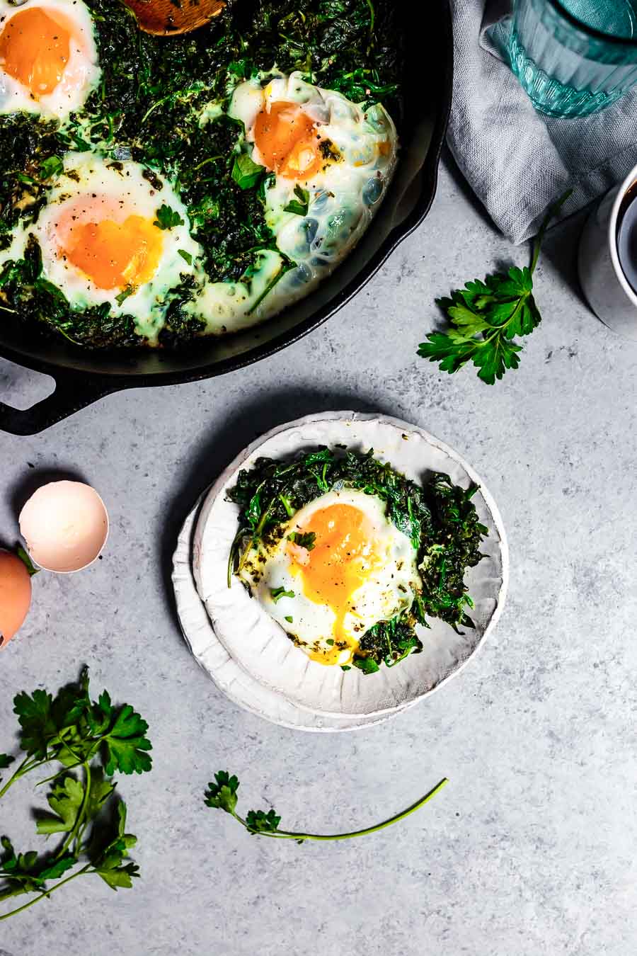 View from above. Just under half of the skillet containing green breakfast shakshuka is visible. nIt is in the top right corner of the image. 
The vegetables are dark green after being cooked and the poached eggs are vibrant inside of the veg.
A portion of green shakshuka with an egg has been put onto a white plate to serve.