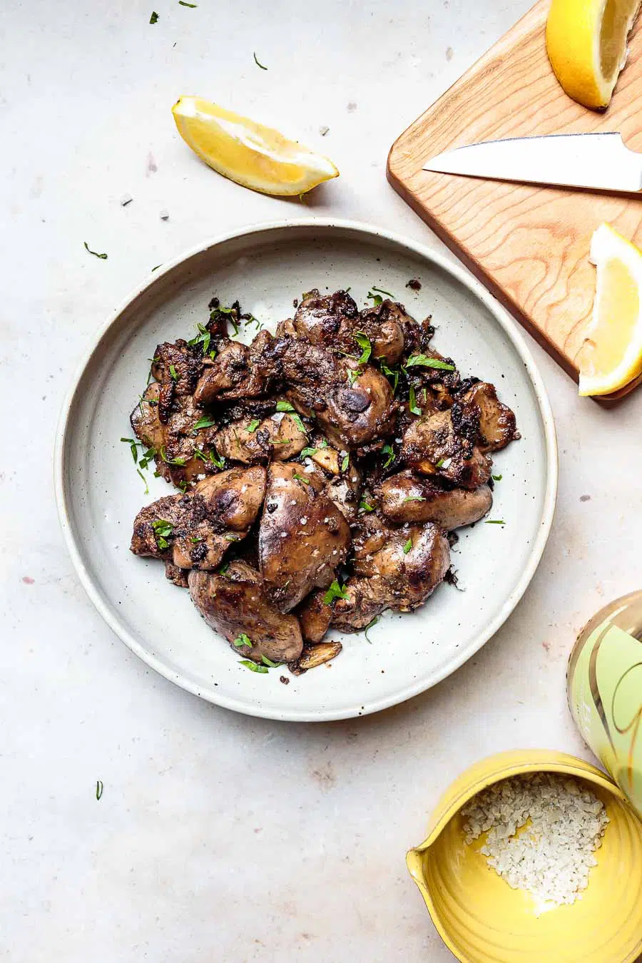 sautéed lemon garlic chicken liver with fresh parsley and lemon wedges on a stoneware plate with a cutting board, knife, and lemon in the background