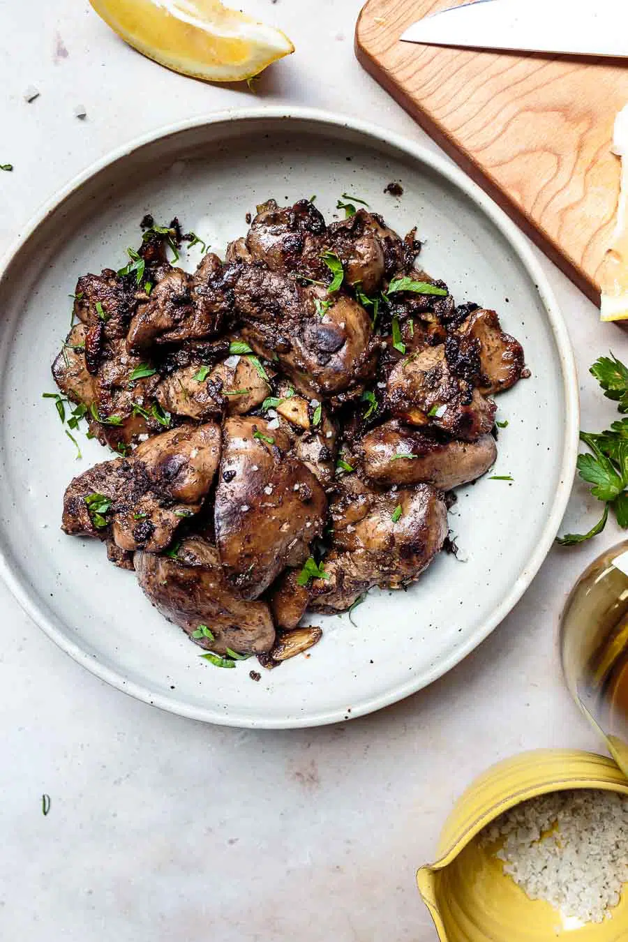 sautéed lemon garlic chicken liver with fresh parsley and lemon wedges on a stoneware plate with a cutting board, knife, and lemon in the background