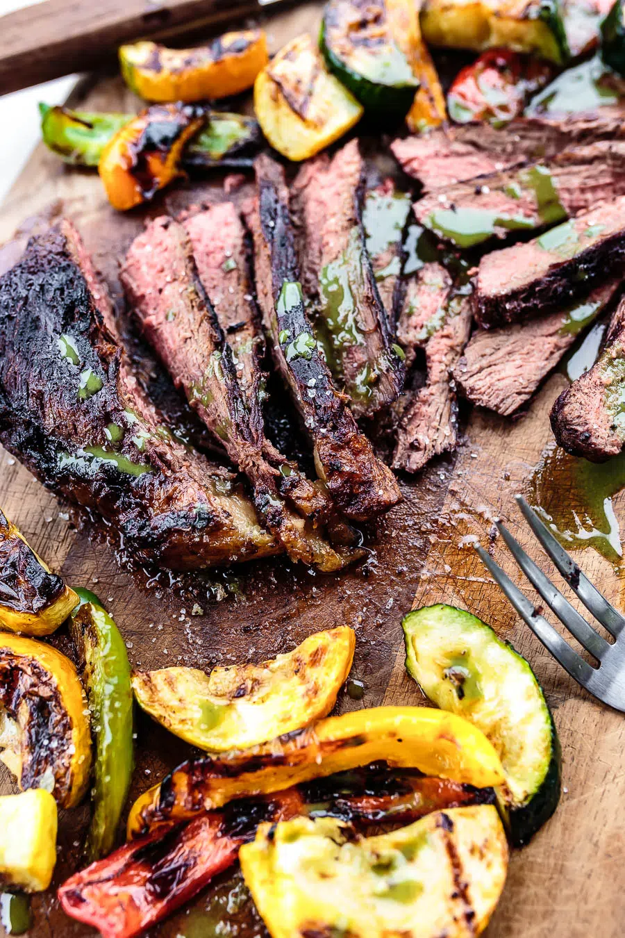 Close up side view of sliced grilled picanha steak with a drizzel of chimichurri on top. Served on a wooden cutting board with grilled vegetables.