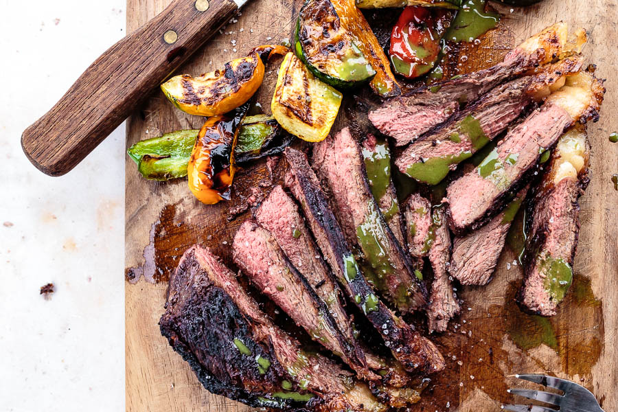 Sliced grilled flank steak with chopped vegetables and avocado dressing served on a wooden cutting board. 