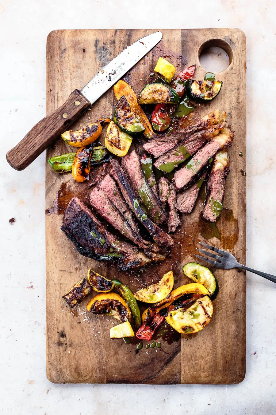 Grilled picanha steak is sliced with chopped vegetables and chimichurri sauce served on a wooden cutting board. A fork and a steak knife are sitting on opposite end of the board.