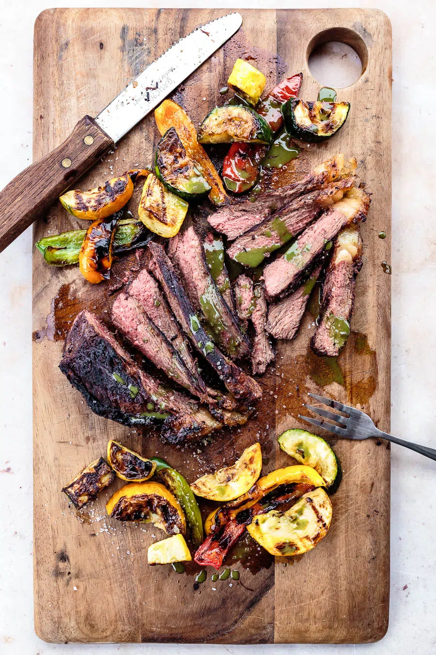 Sliced grilled picanha steak with roasted vegetables and chimichurri sauce served on a wooden cutting board. A fork and a steak knife are sitting on opposite end of the board.