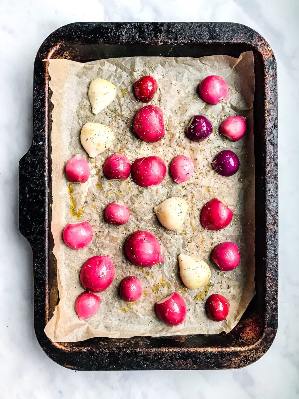 radishes that have been sliced in half and tossed in olive oil, salt, and dried rosemary resting on a prepared baking sheet ready for the oven