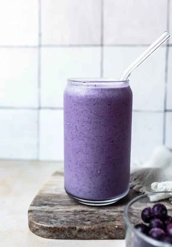 Glass with purple blueberry bone broth smoothie in it, sitting on a wooden chopping board.