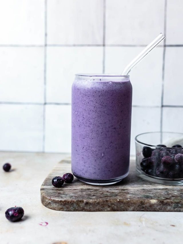 An egg free breakfast - In a white kitchen, a glass of blueberry and cardamon bone broth smoothie is in a clear tall glass glass on a marble board. There are extra blueberries in a glass bowl next to it and scattered around the kitchen.