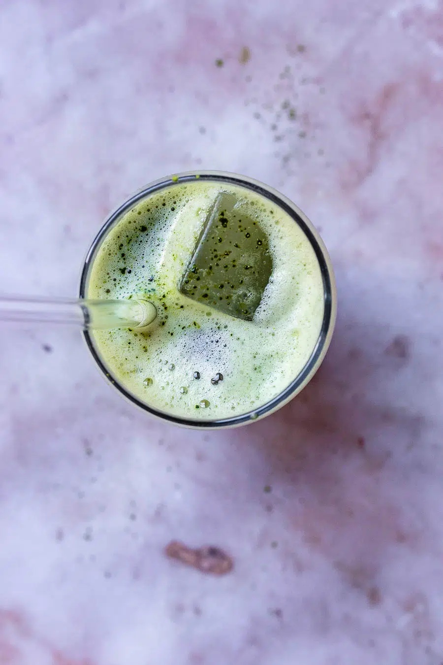 Top view of a purple work top with a glass filled with iced matcha and ice, with a clear straw in it.