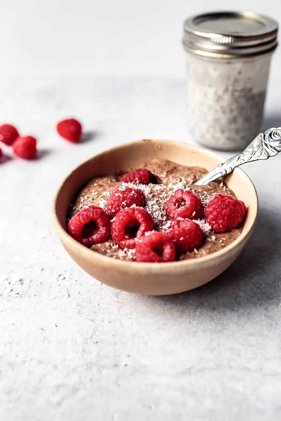 Blended chia pudding in a clay bowl on a white counter top. The pudding is topped with raspberries and desiccated coconut. There is a silver spoon sitting inside the bowl and a few scattered raspberries and a jar of soaking chia seeds on the counter top.