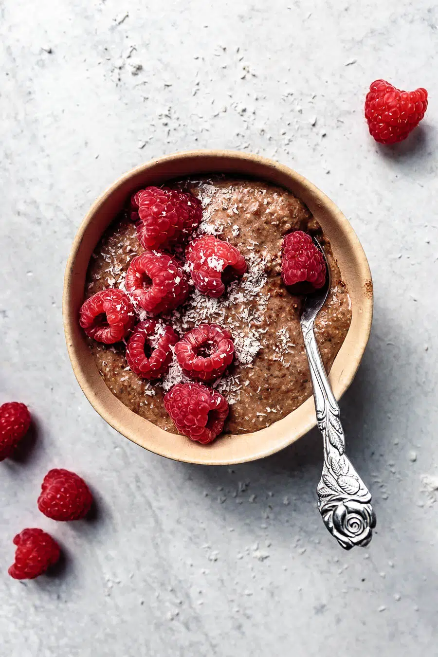 Birds eye view of blended chocolate chia pudding in a clay bowl on a white counter top. The pudding is topped with raspberries and desiccated coconut. There is a silver spoon sitting inside the bowl and 2 stray raspberries on the counter top.