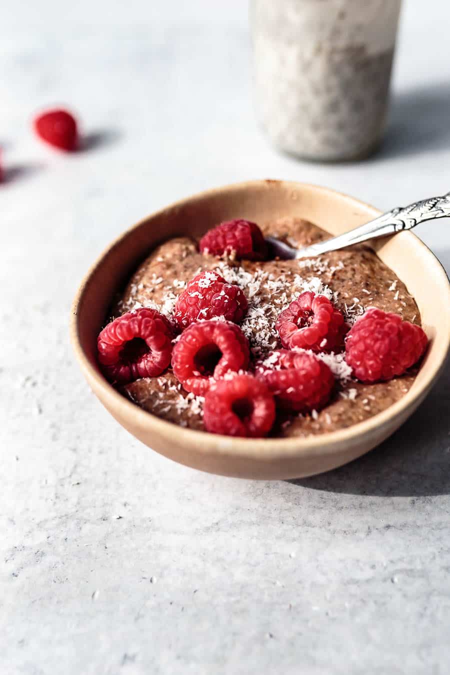 Blended chocolate chia pudding in a clay bowl on a white counter top. The pudding is topped with raspberries and desiccated coconut. There is a silver spoon sitting inside the bowl a jar of soaking chia seeds on the counter top.