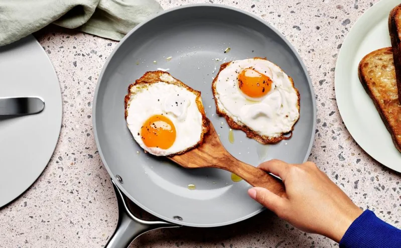 On a white marbled counter top, 1 of the grey healthy nonstick cookware pans is sitting. Someone is using a wooden spoon to easily pick up a fried egg.