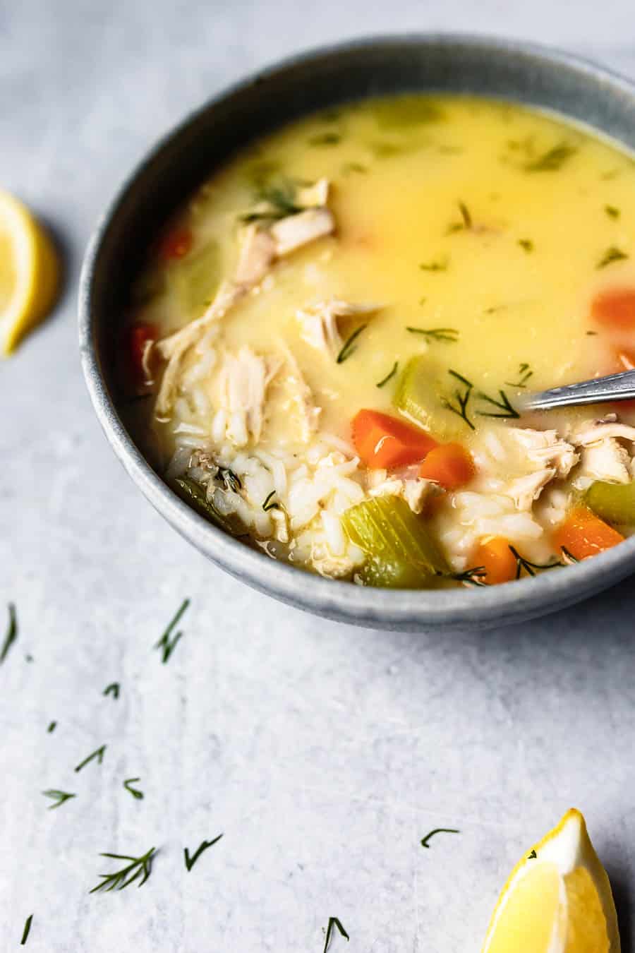 Partial view of this Lemon Chicken Avgolemono Soup. It is a creamy yellow color and has carrots, celery, chicken and rice inside it. It has been garnished with dill and served in a grey bowl. It is place on top of a grey counter where there is leftover lemon wedges laying.