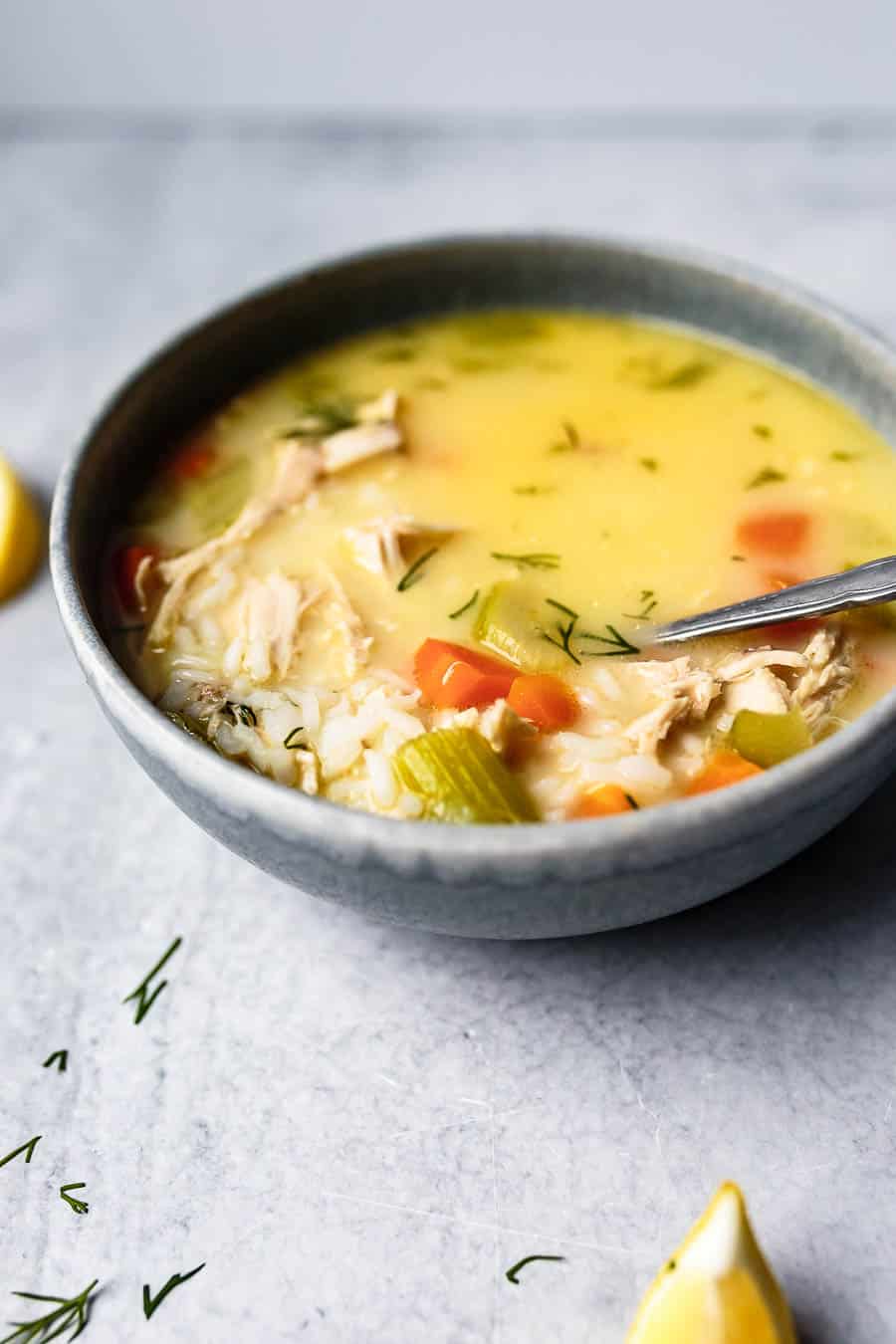 Yellow color Avgolemono Soup in a dark grey bowl, sitting on a light grey table. There is a silver spoon in the bowl and it is full of vegetables like carrot and celery. There is also rice and chicken in the soup.