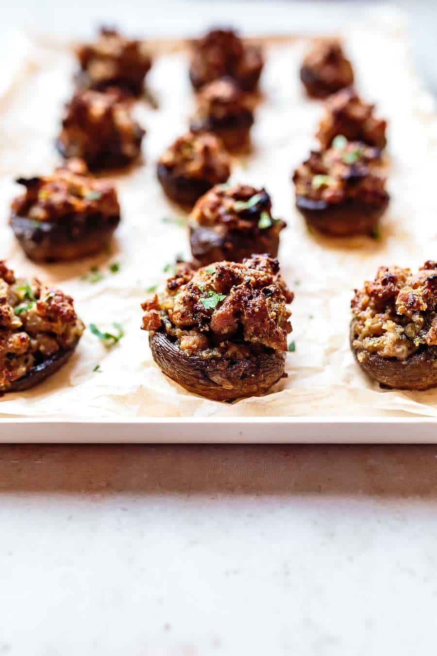 A slight angle, low perspective. A baking tray lined with baking paper and topped with lots of baked sausage stuffed mushrooms. The mushrooms closest to the camera are in focus and moving back, they decrease in focus.