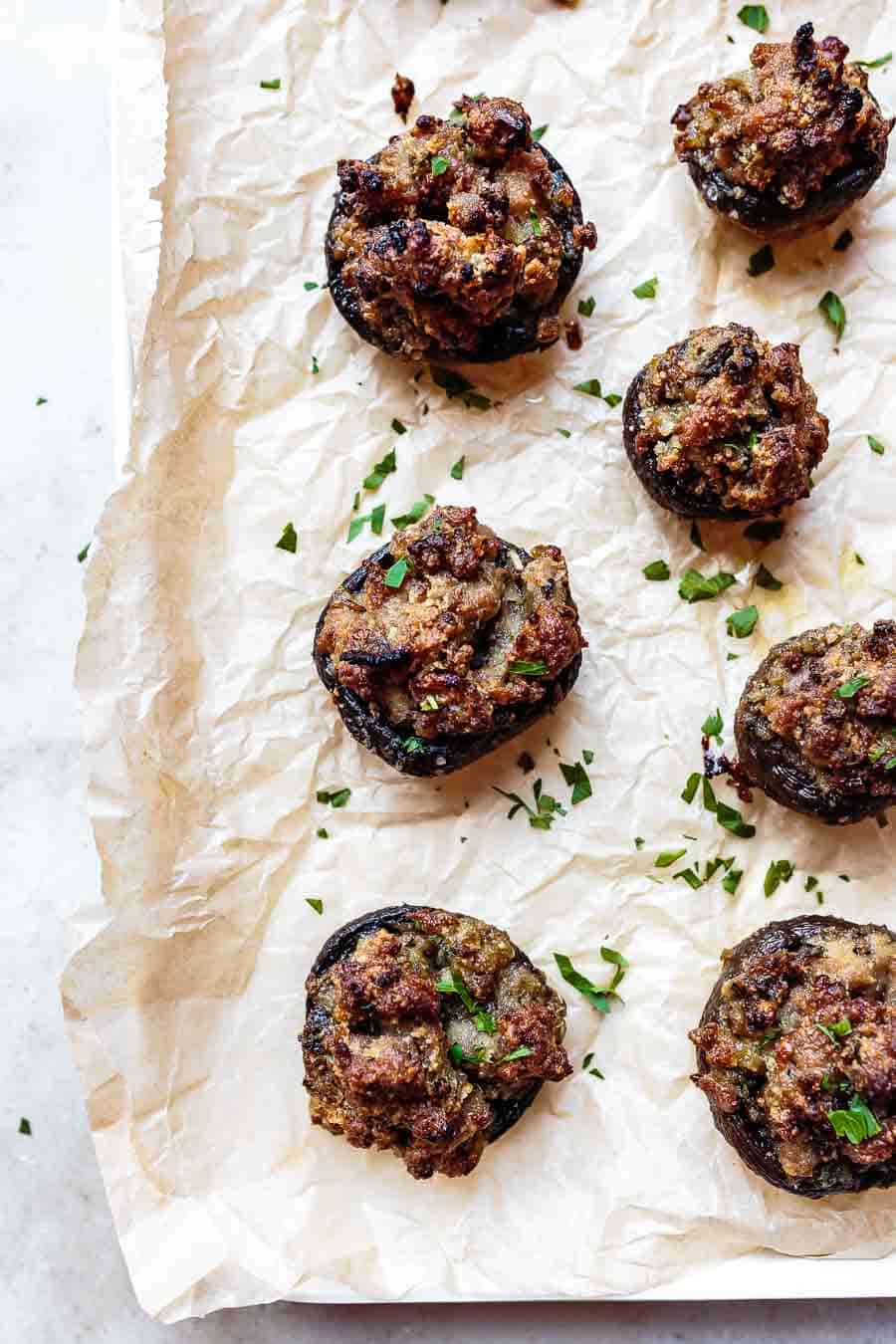 View from above of 7 baked sausage stuffed mushrooms sitting on a piece of parchment paper. The mushroom cap is facing down and they have been garnished with some fresh herbs.